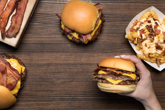 Bacon Double Cheeseburger held in hand with other bacon cheeseburger and bacon fries in the background