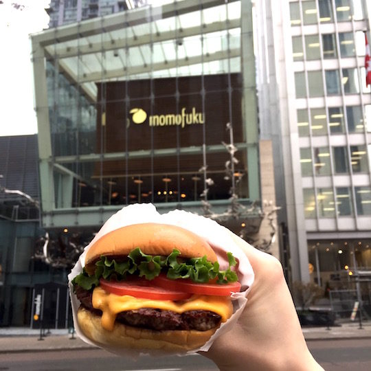 shake shack burger being held by a hand with momofuku store in the background