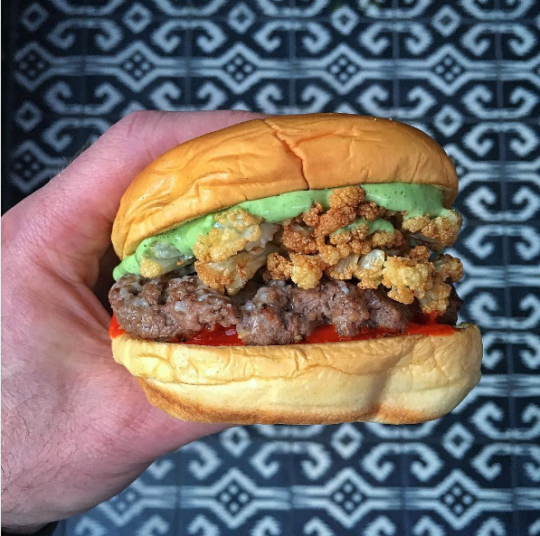 Shake Shack and Chef Lee Wolen's BOKA inspired burger held in hand in front of print background