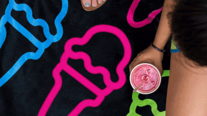 black towel with green, blue, and pink ice cream outlines repeating on it. Two feet from one person are seen at the top. On the side, part of another person can be seen, holding a shake.