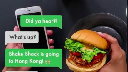 There is a picture of a hand holding a phone, one with a burger, and a drink next to it, pictured from above. Over it are texting bubbles: Did ya hear?! What's up? Shake Shack is going to Hong Kong. OMG.