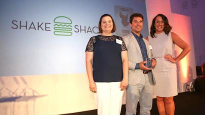 three people with nametags on standing in front of a wall with the shake shack logo on it