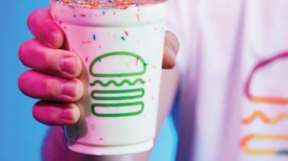 Person wearing a shirt with a rainbow shake shack logo, holding a shake with rainbow sprinkles in it.